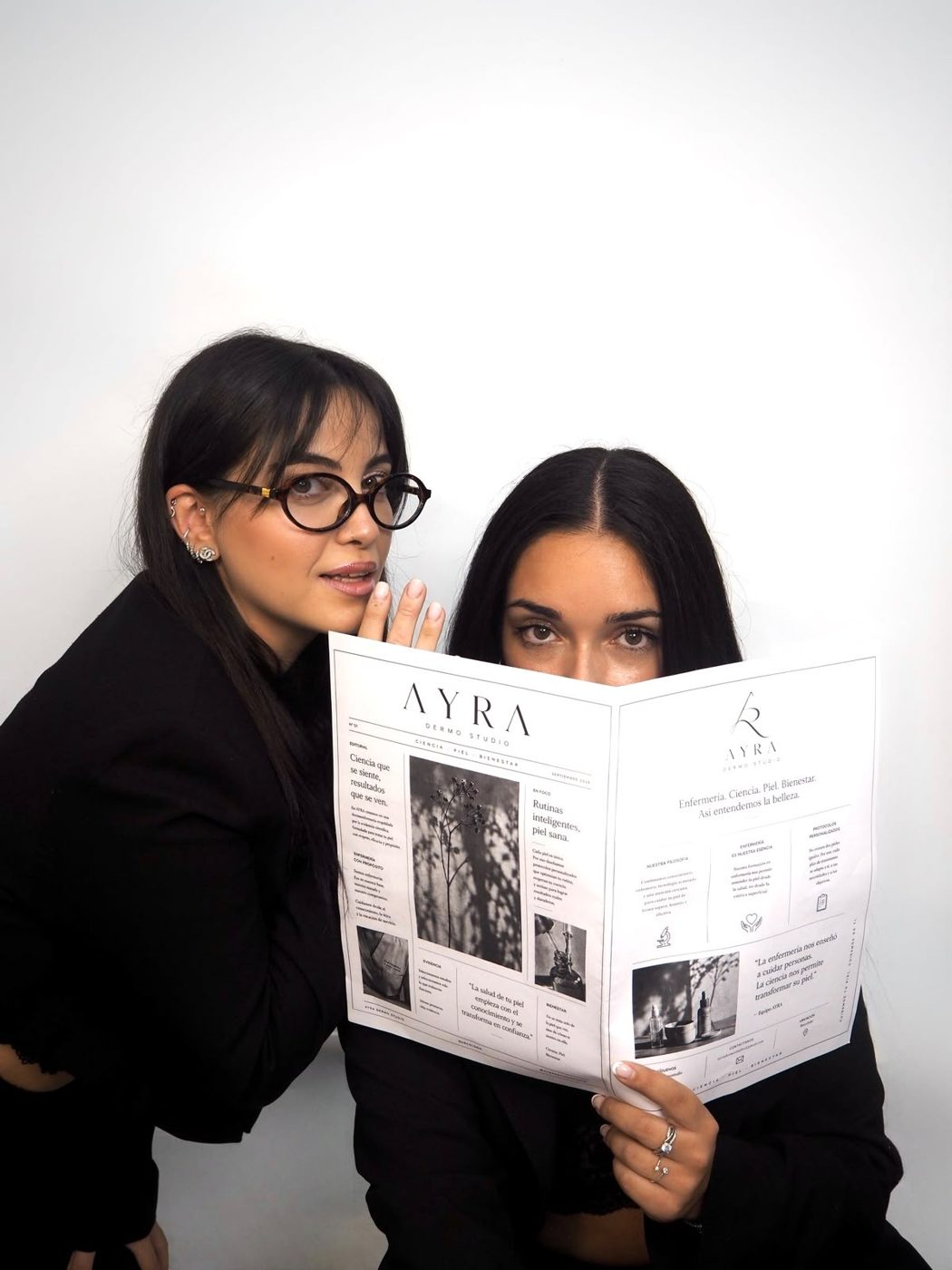 Alba, con gafas, susurra a Raquel, que se tapa media cara con el periódico AYRA